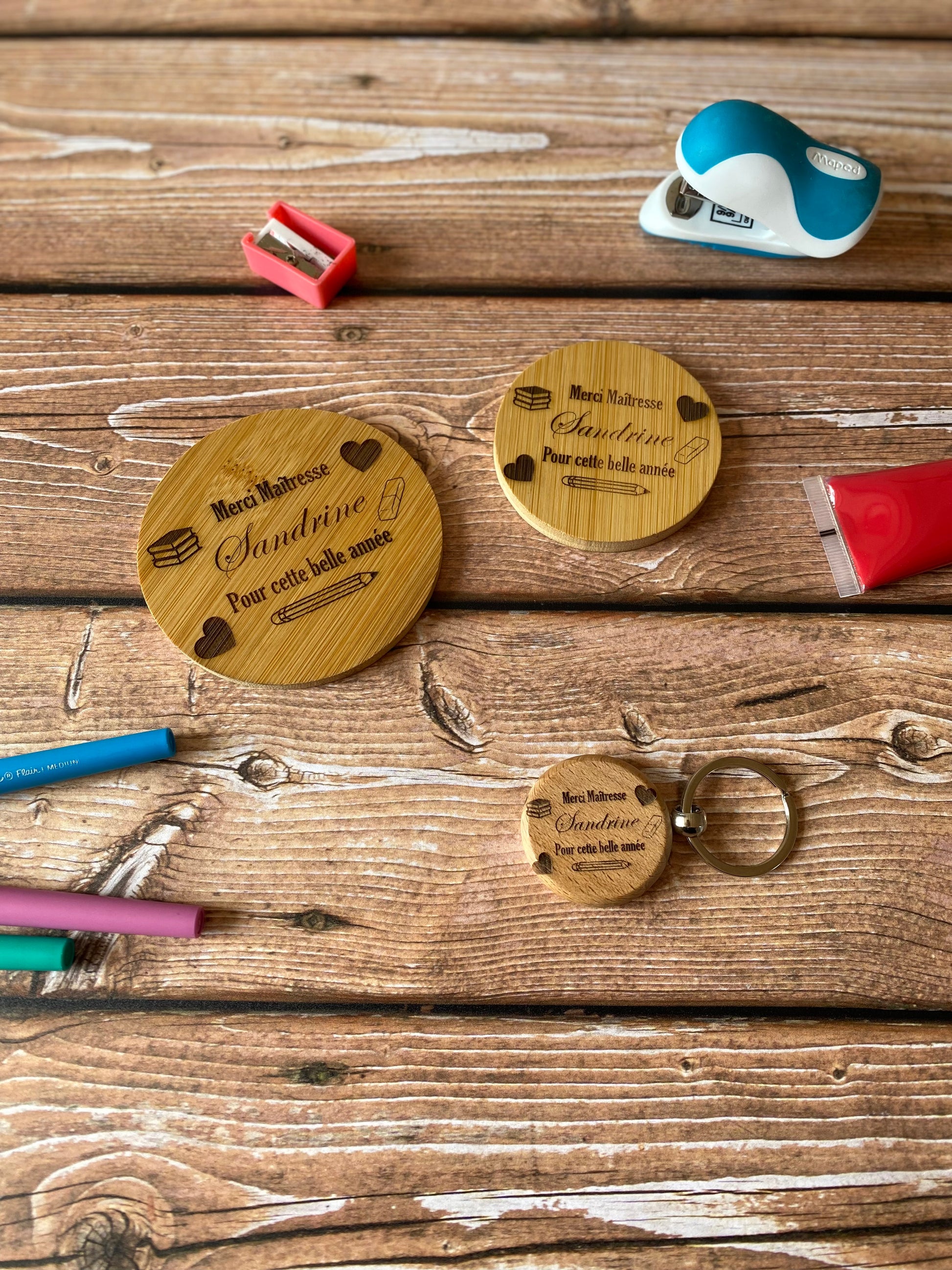 Pack cadeau personnalisé pour enseignant(e) avec miroir de poche, porte-clé et dessous de verre en bambou gravés, disposés sur un bureau d'écolier avec des fournitures scolaires autour